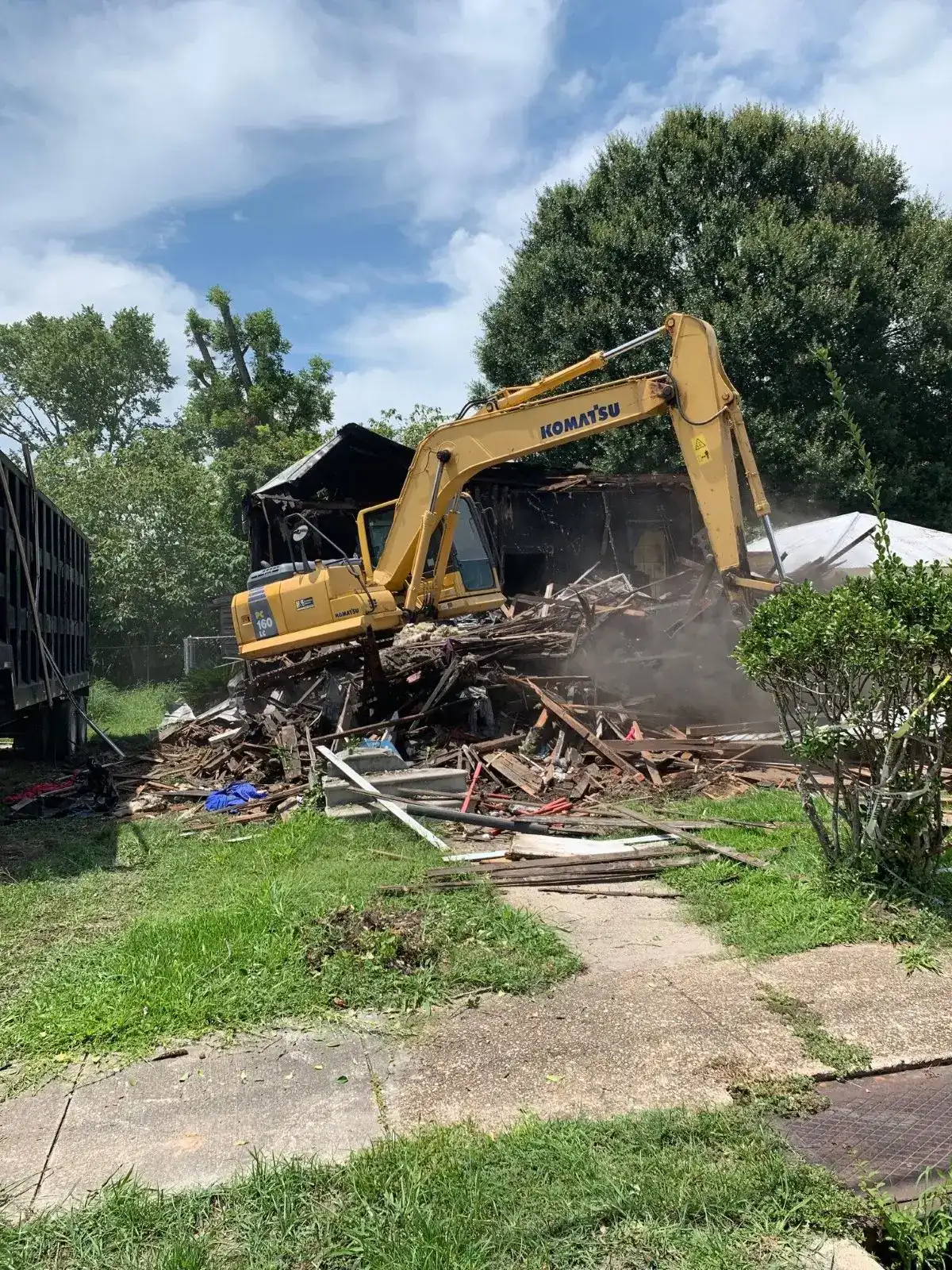 Excavator demolishing a house surrounded by trees and grass.